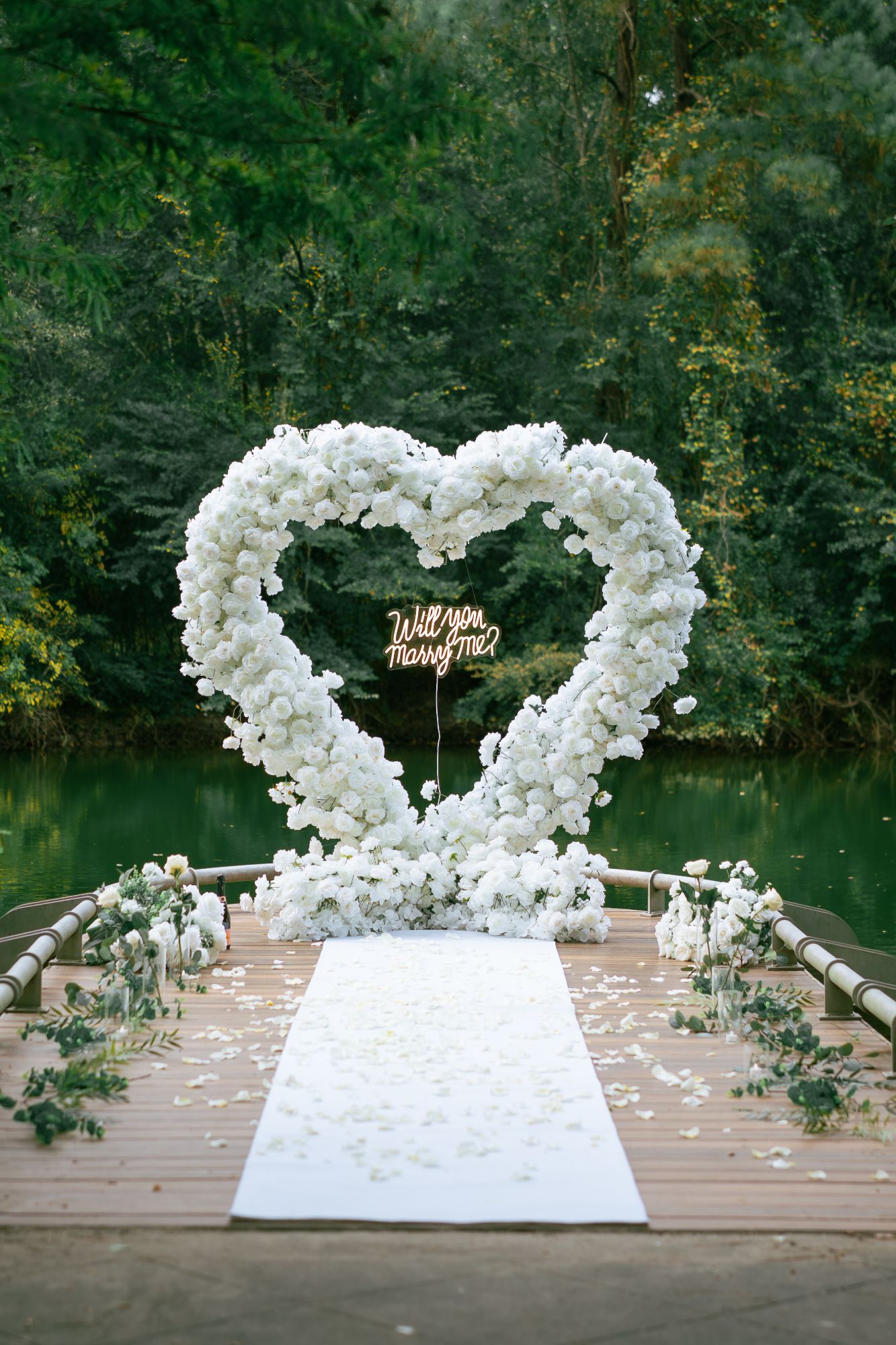 White rose floral heart arch with “Will you marry me?” neon sign, white carpet and rose petal path on a lakeside dock